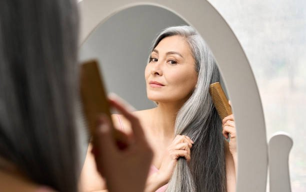 An Asian woman looking in an oval-shaped mirror combing her long gray hair.