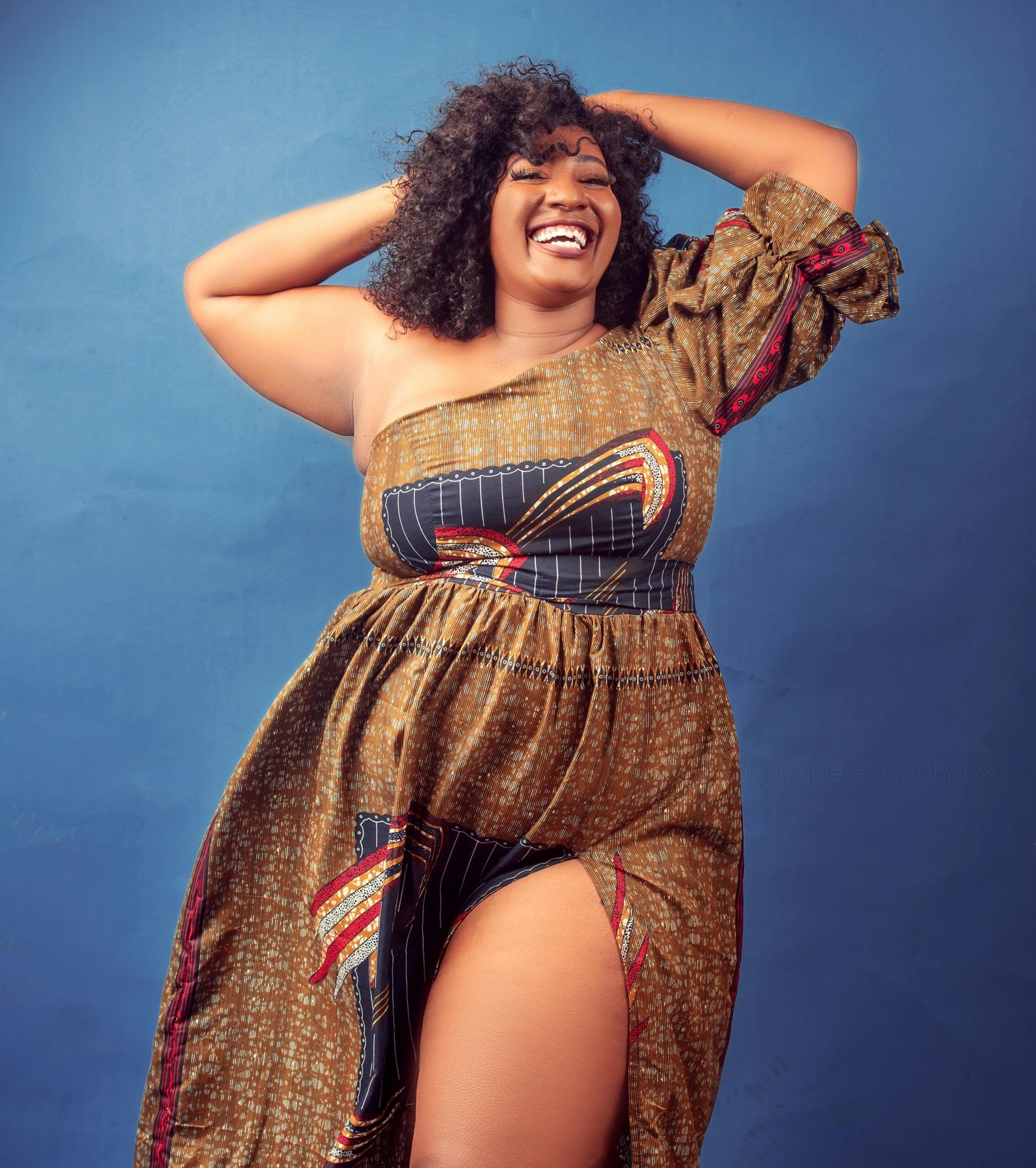 A plus-size African American woman with shoulder-length curly hair wearing a long, african-print dress. The dress has a one-shoulder, puffy sleeve and a thigh-high split. She is smiling while lifting her hands and arms which are touching the back of her head.