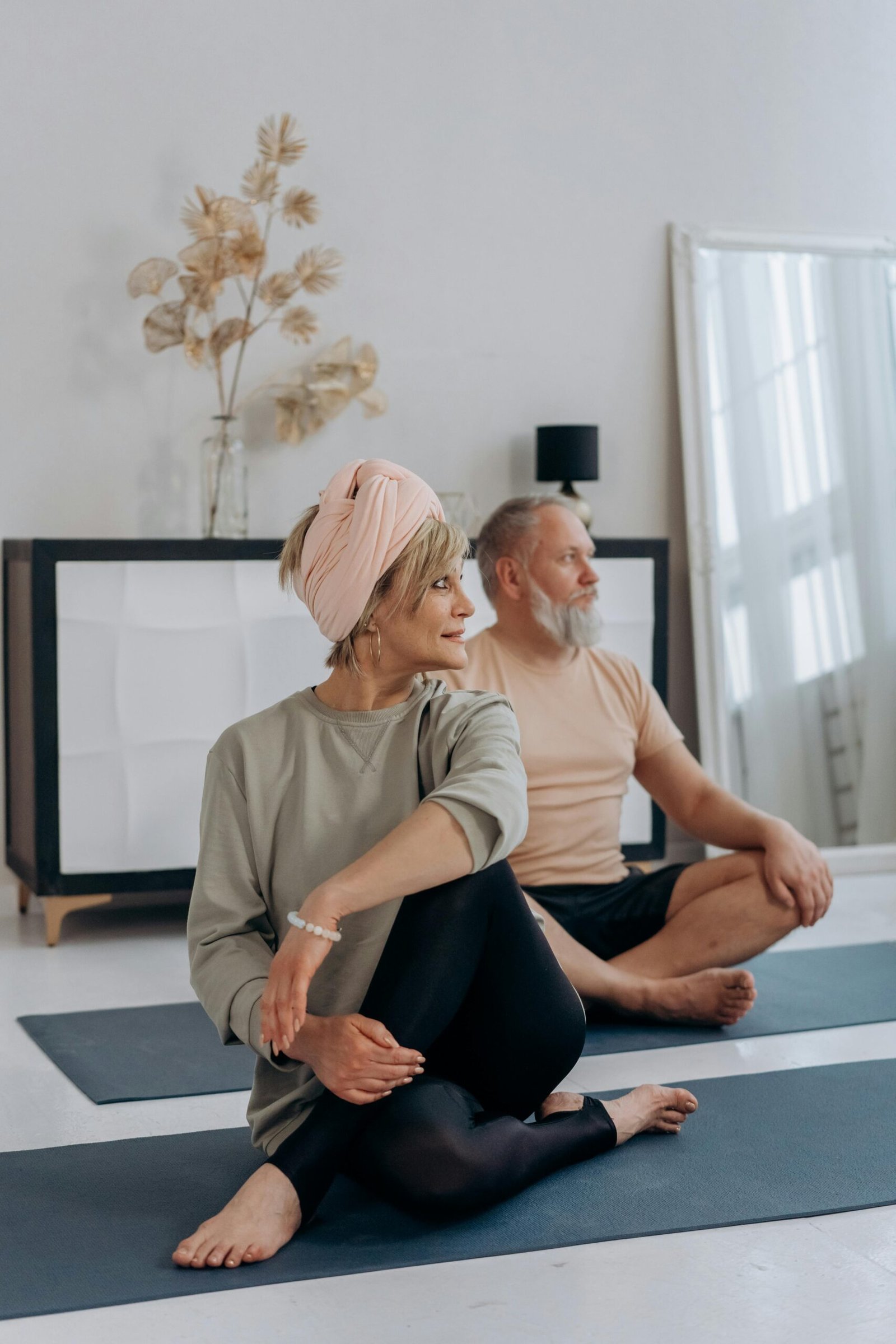 A Caucasian lady sitting on the floor of her home with her husband in back of her, both in a yoga position on floor mats.